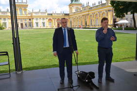 Pokaż zdjęcie: Dwóch mężczyzn, jeden stoi z psem i przemawia, drugi obok wykonuje tłumaczenie na język migowy